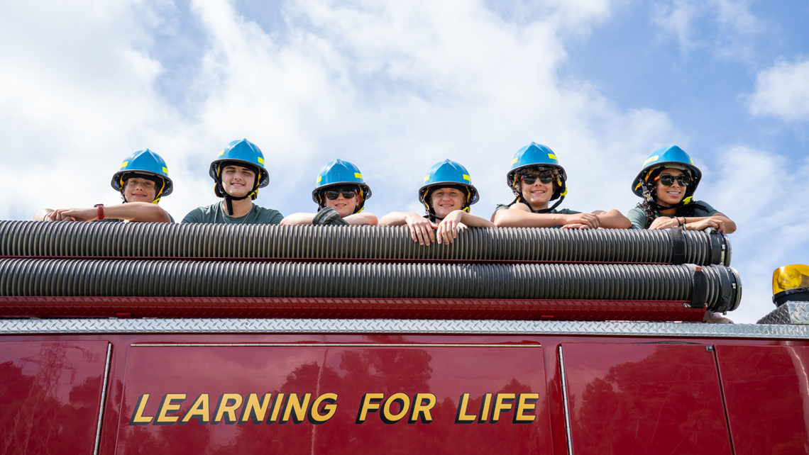 Campers in blue firefighting helmets standing on fire truck. Deep blue sky in background.