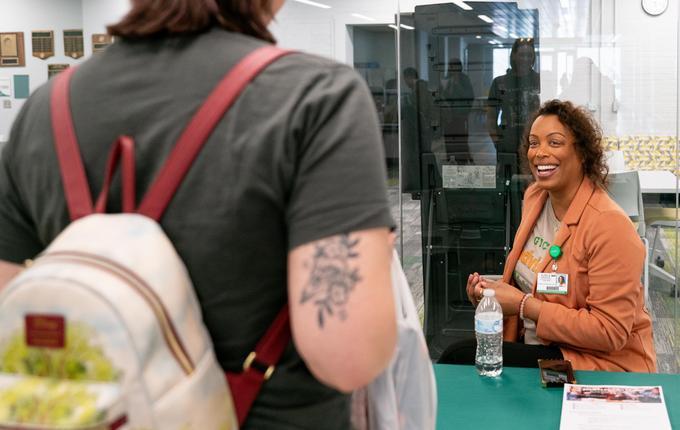 Looking from behind a student wearing a backpack and who has an arm tattoo, a black female counselor smiles wearing a orange brown jacket and gray GTCC T-shirt.