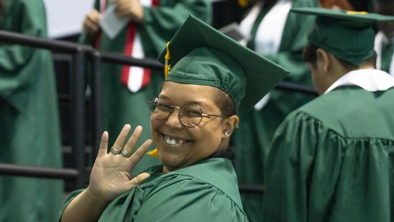 A female student smiles and waves during commencement.