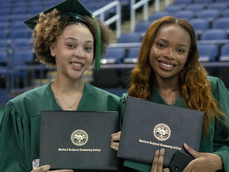 Two female students smile and hold their diploma covers.