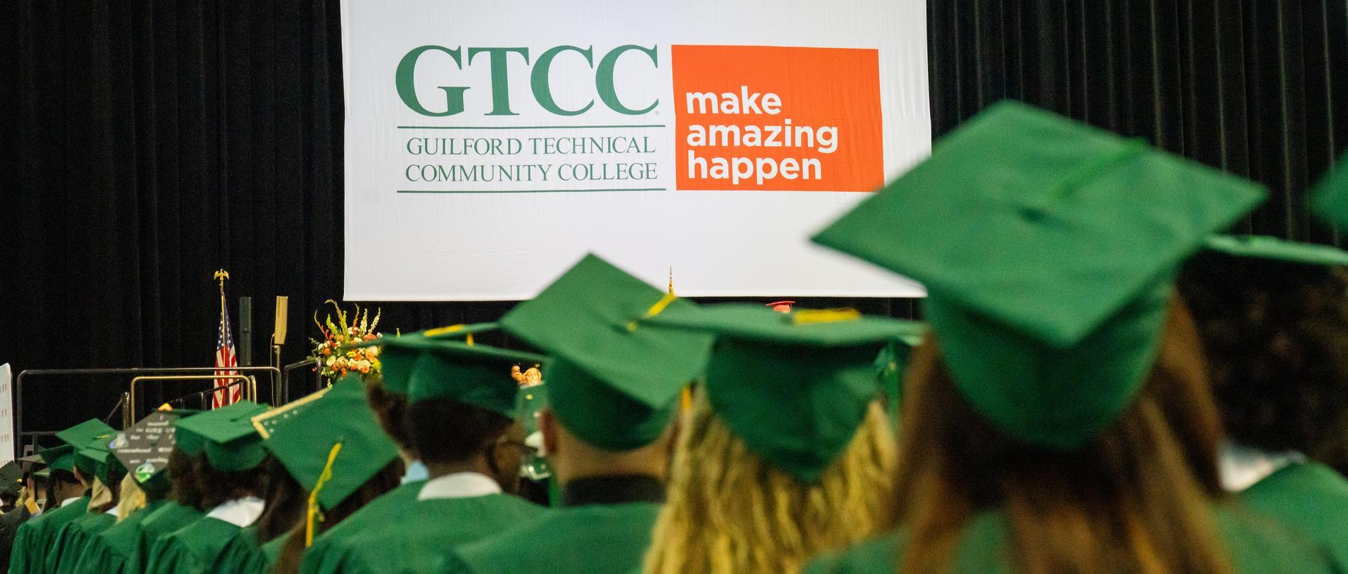 GTCC graduates in green caps and gowns face the stage during commencement, with a large GTCC “Make Amazing Happen” banner in view.