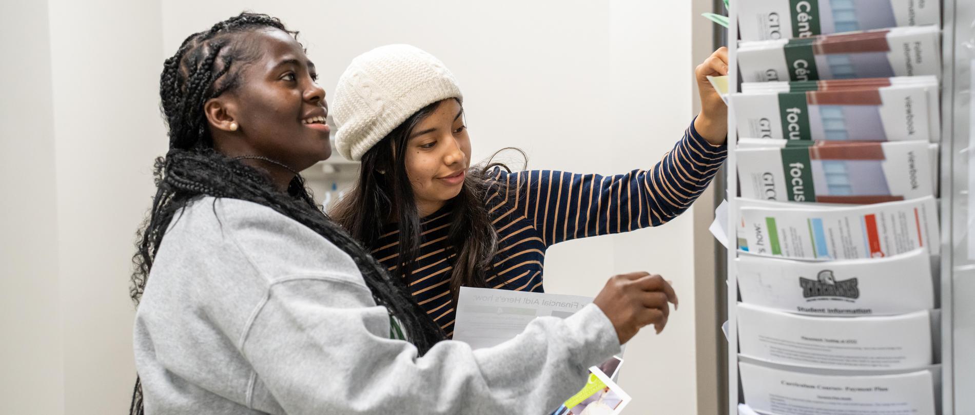 Two GTCC students smile while browsing scholarship and student resource brochures from a rotating display rack.