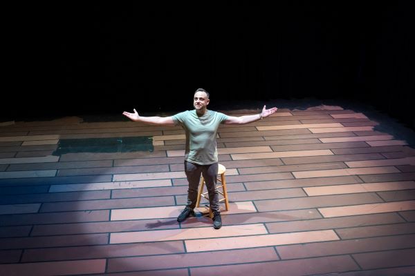 Nick Barbato stands on stage with outstretched arms with featured lighting highlighting him.