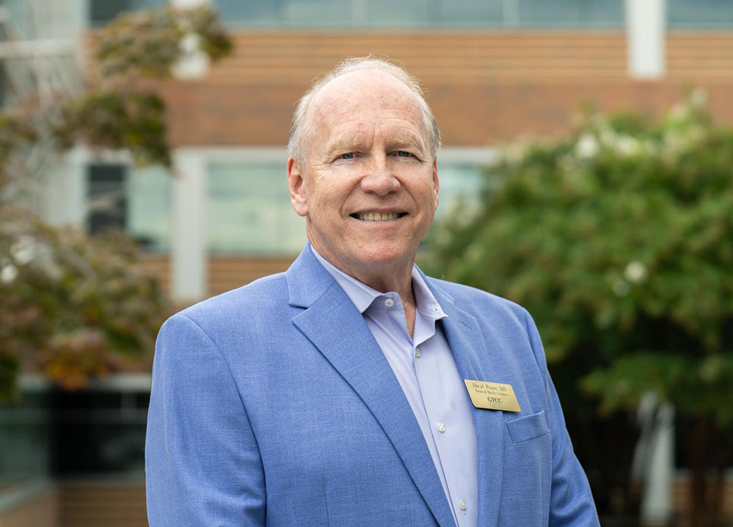 David Frazee, wearing a light blue blazer and GTCC name badge, smiles while standing outdoors in front of a campus building with trees in the background.