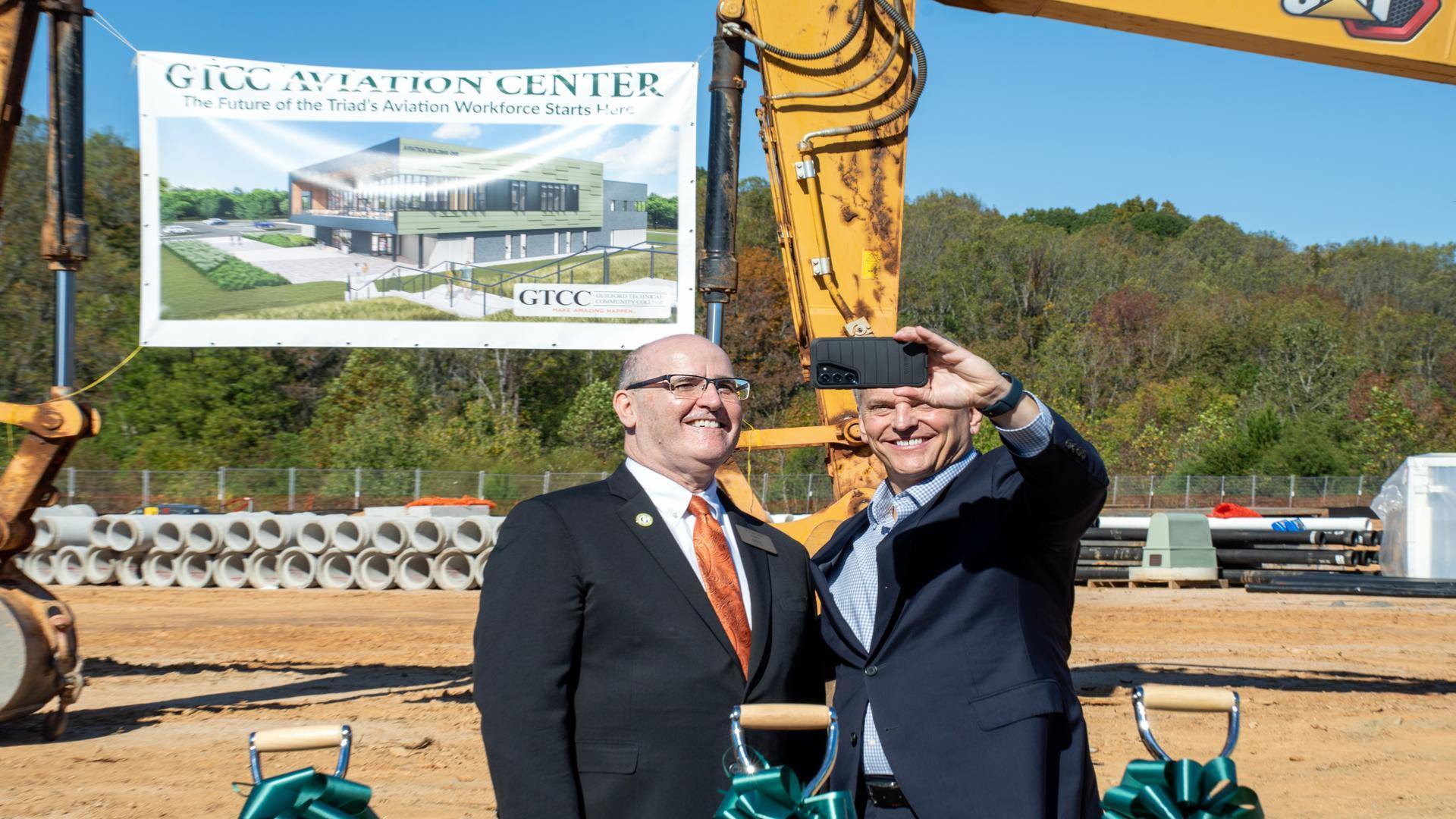 GTCC President Dr. Anthony Clarke and North Carolina Governor Josh Stein take a selfie together during the Aviation Center groundbreaking ceremony, with a rendering of the new facility displayed behind them.