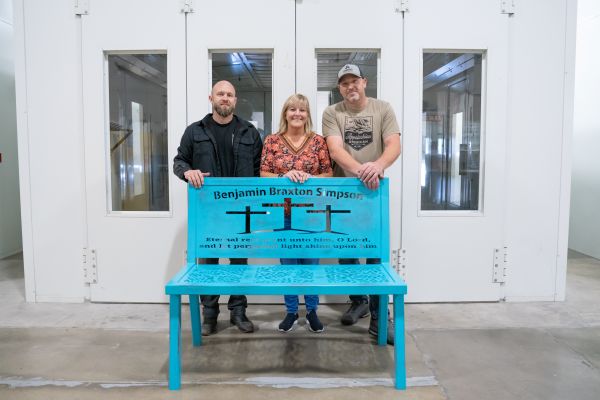 Three people stand behind a custom teal memorial bench inside a GTCC workshop, featuring the name “Benjamin Braxton Simpson” and a cross design on the backrest.