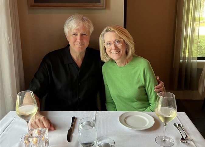 Jim and Deborah Hooper sit together at a restaurant table, smiling for the camera with glasses of white wine and place settings in front of them.